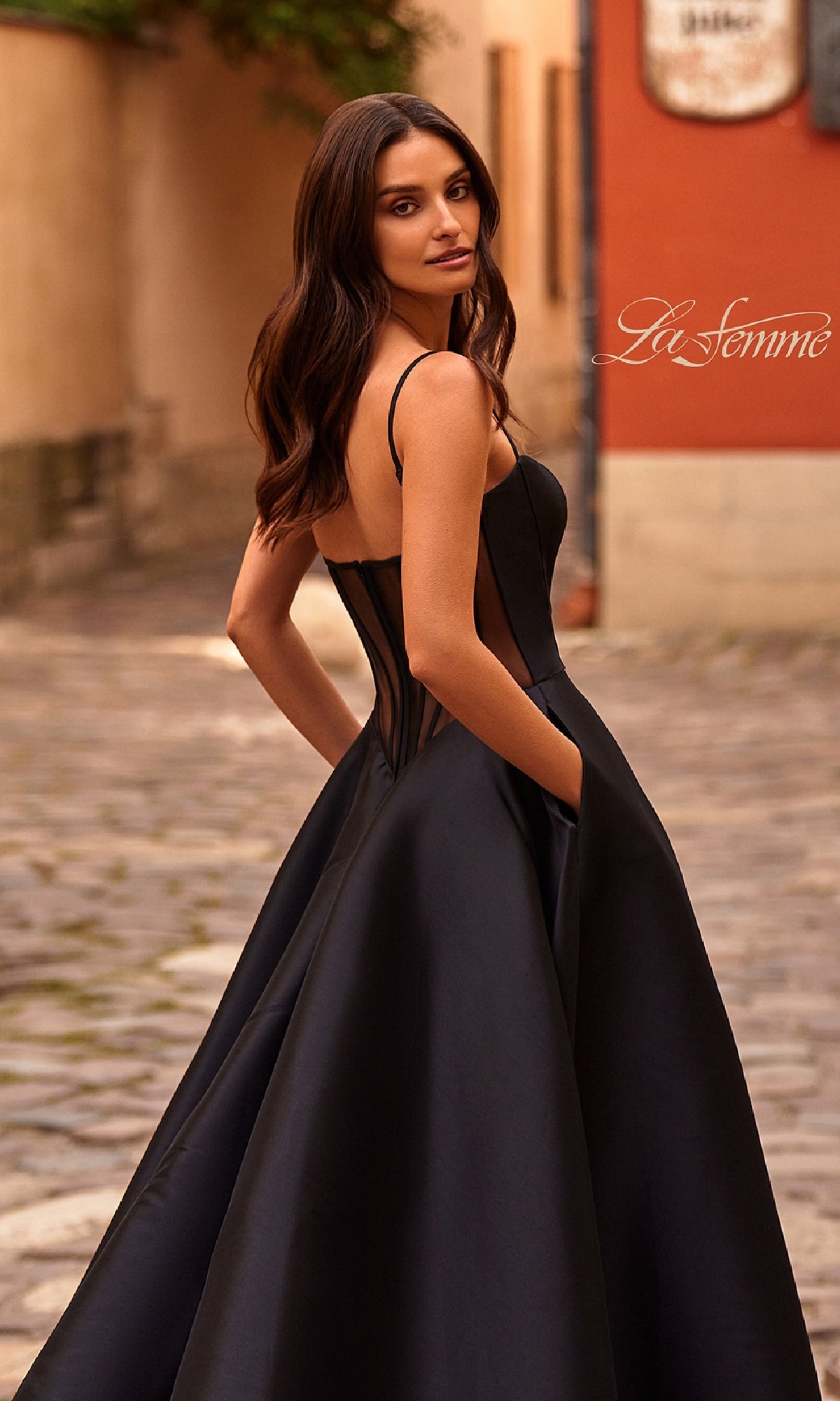 A woman in a Sheer-Corset Long A-Line Prom Gown (La Femme 32670) stands on a cobblestone street, glancing over her shoulder as